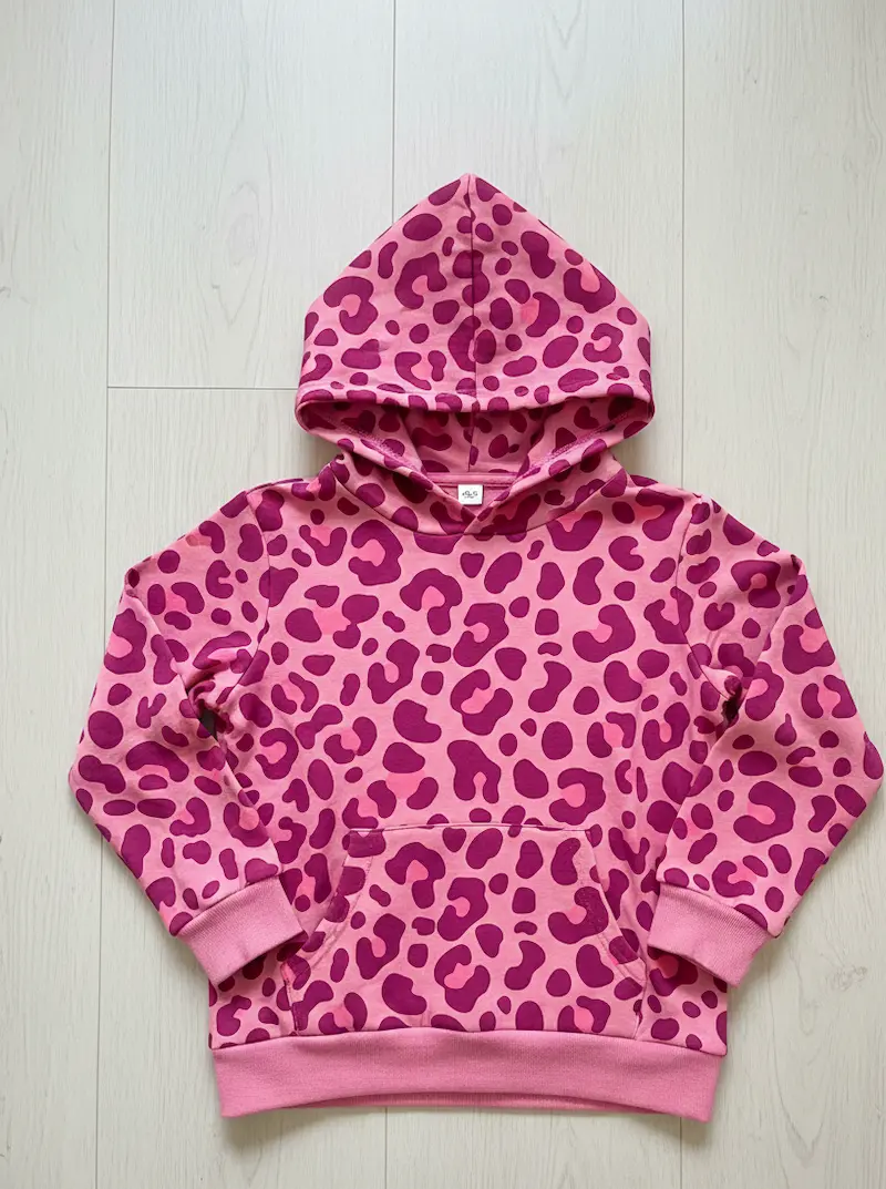Ein flach liegender Kinder-Kapuzenpullover (Hoodie) mit Kängurutasche, genäht aus elastischer Baumwolljersey-Meterware. Der Stoff zeigt ein Allover-Leopardenmuster in kräftigen Pink-, Beeren- und Rosatönen auf einem hellen Holzboden.