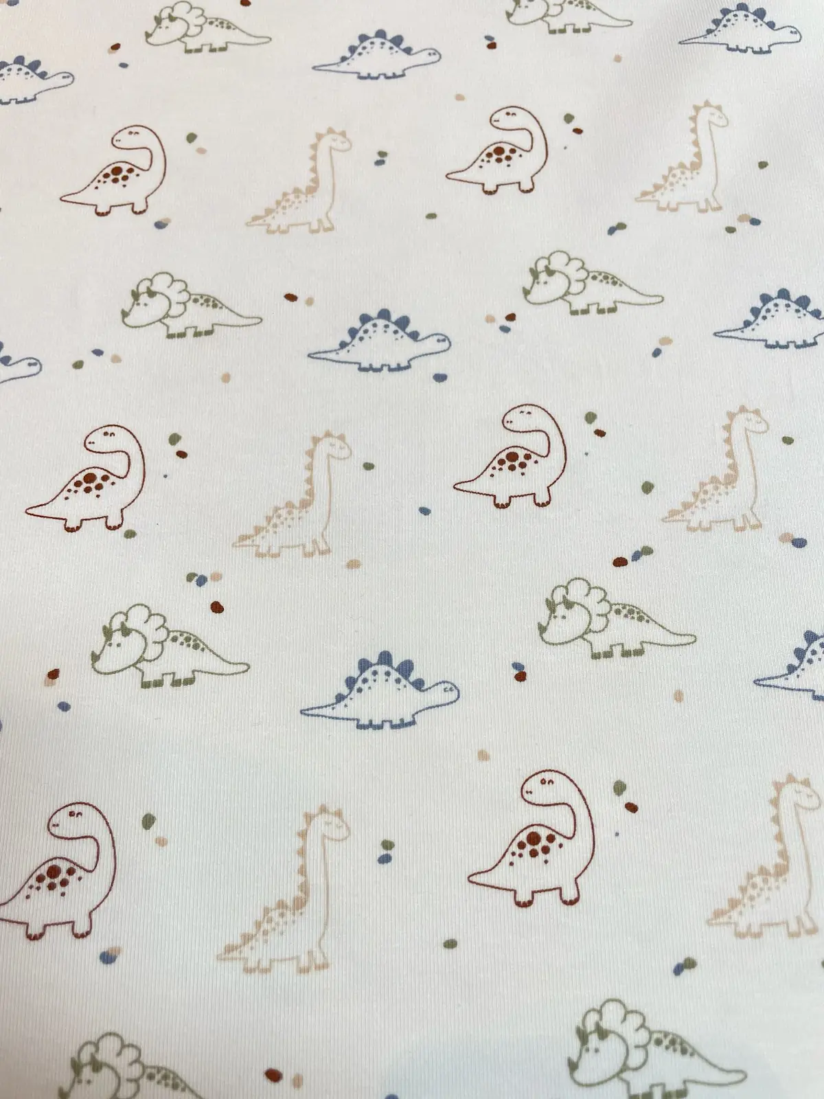 Weißer Jersey-Stoff mit kleinen Dinosaurier-Motiven wie Triceratops und Stegosaurus in Blau, Rostbraun und Beige mit bunten Sprenkeln.