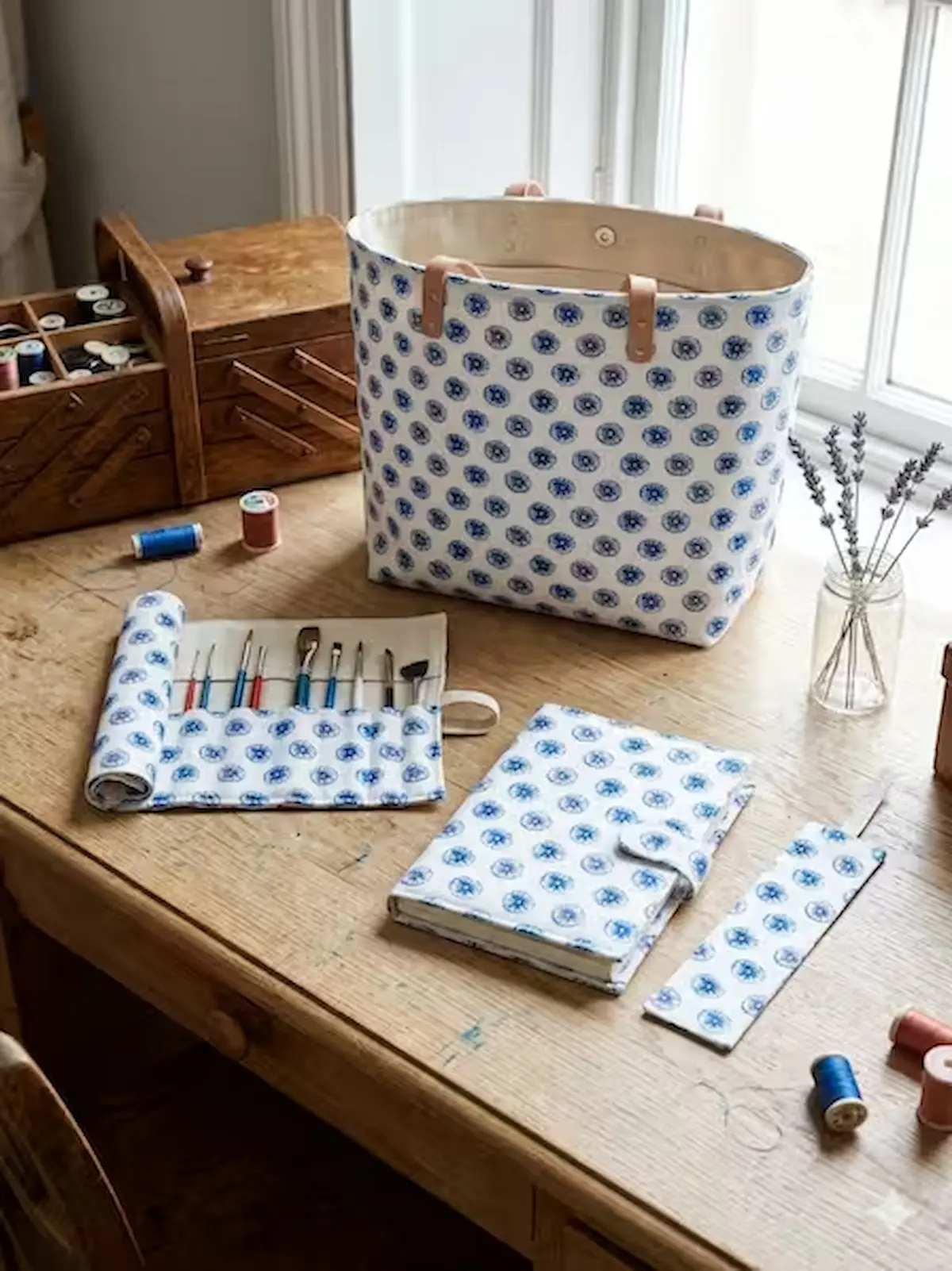 Genähte Accessoires aus weißem Baumwoll-Webware-Stoff mit einem blauen, kreisförmigen Blütenmuster auf einem Holztisch. Zu sehen sind eine große Einkaufstasche (Shopper) mit Lederhenkeln, eine ausgerollte Pinselrolle mit Pinseln, eine passende Buchhülle m