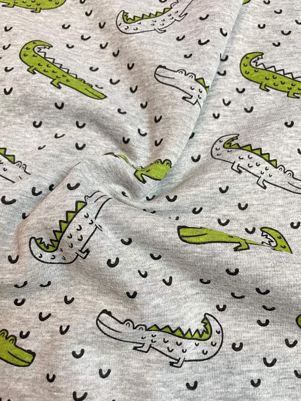 Detailaufnahme eines weichen Alpenfleece-Stoffes in Grau-Melange mit Krokodil-Motiven. Die Stofffalten zeigen die geschmeidige, schwere Qualität des Materials für Winterbekleidung.