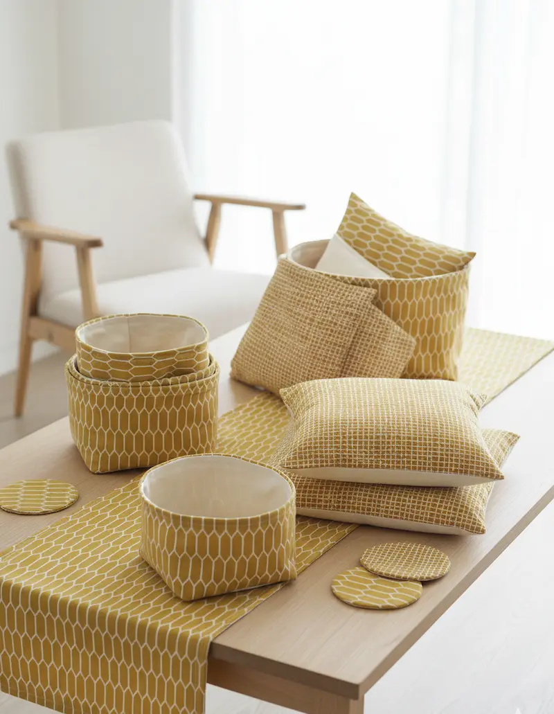 Verschiedene selbstgenähte Wohnaccessoires aus senfgelber Baumwoll-Webware mit geometrischem Wabenmuster auf einem Holztisch: Ein langer Tischläufer, Utensilo-Körbchen, Dekokissen und runde Untersetzer im Scandi-Stil.