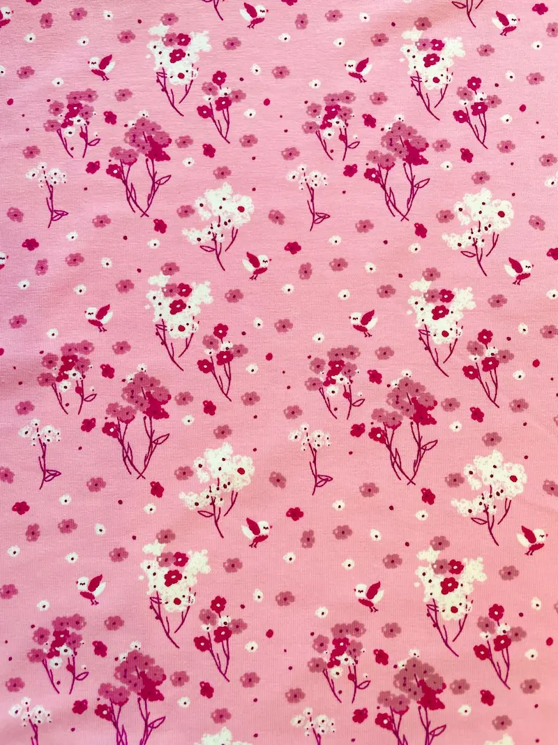 Detaillierte Nahaufnahme von hellrosa Baumwolljersey als Meterware mit einem verspielten Allover-Print aus kleinen, fliegenden Vögeln in Pink und Weiß sowie zarten Frühlingsblumen und Blütenzweigen.