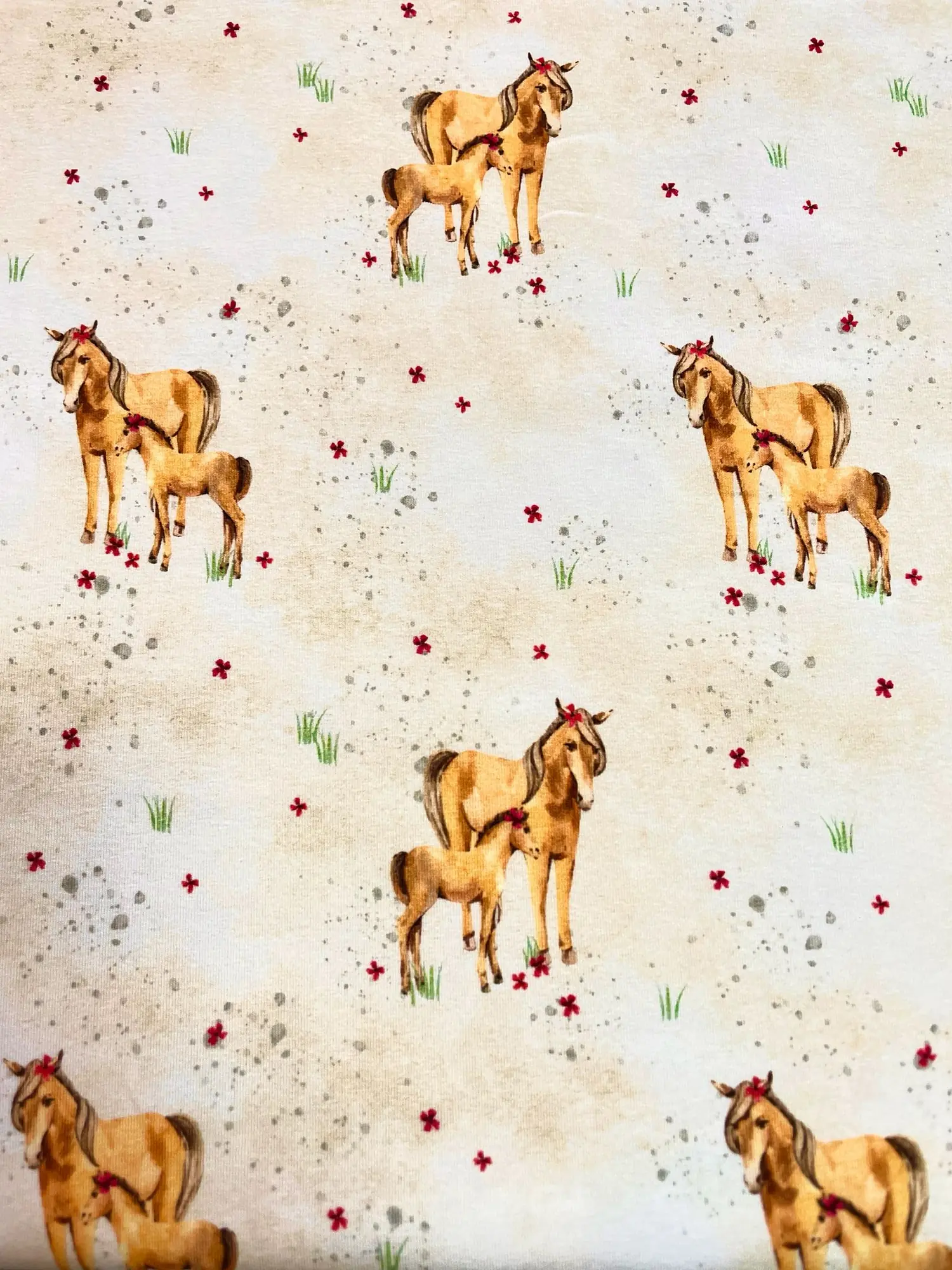 Jersey-Stoff auf hellem, cremefarbenem Hintergrund mit sanften beige-braunen Schattierungen. Das Muster zeigt eine Stute mit ihrem Fohlen, beide mit kleinen roten Blumen im Haar, umgeben von zarten Gräsern, roten Blüten und grauen Pünktchen im Wasserfarbe