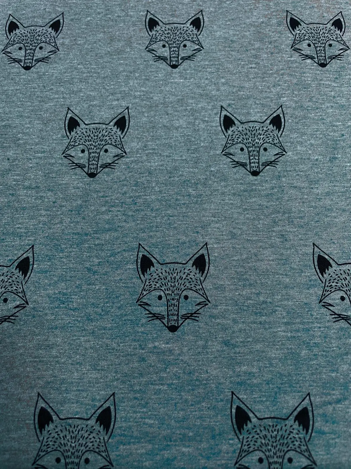Flach liegender, petrolgrün melierter Jerseystoff als Meterware, bedruckt mit einem minimalistischen Allover-Muster aus schwarz gezeichneten Fuchsgesichtern im grafischen Stil.