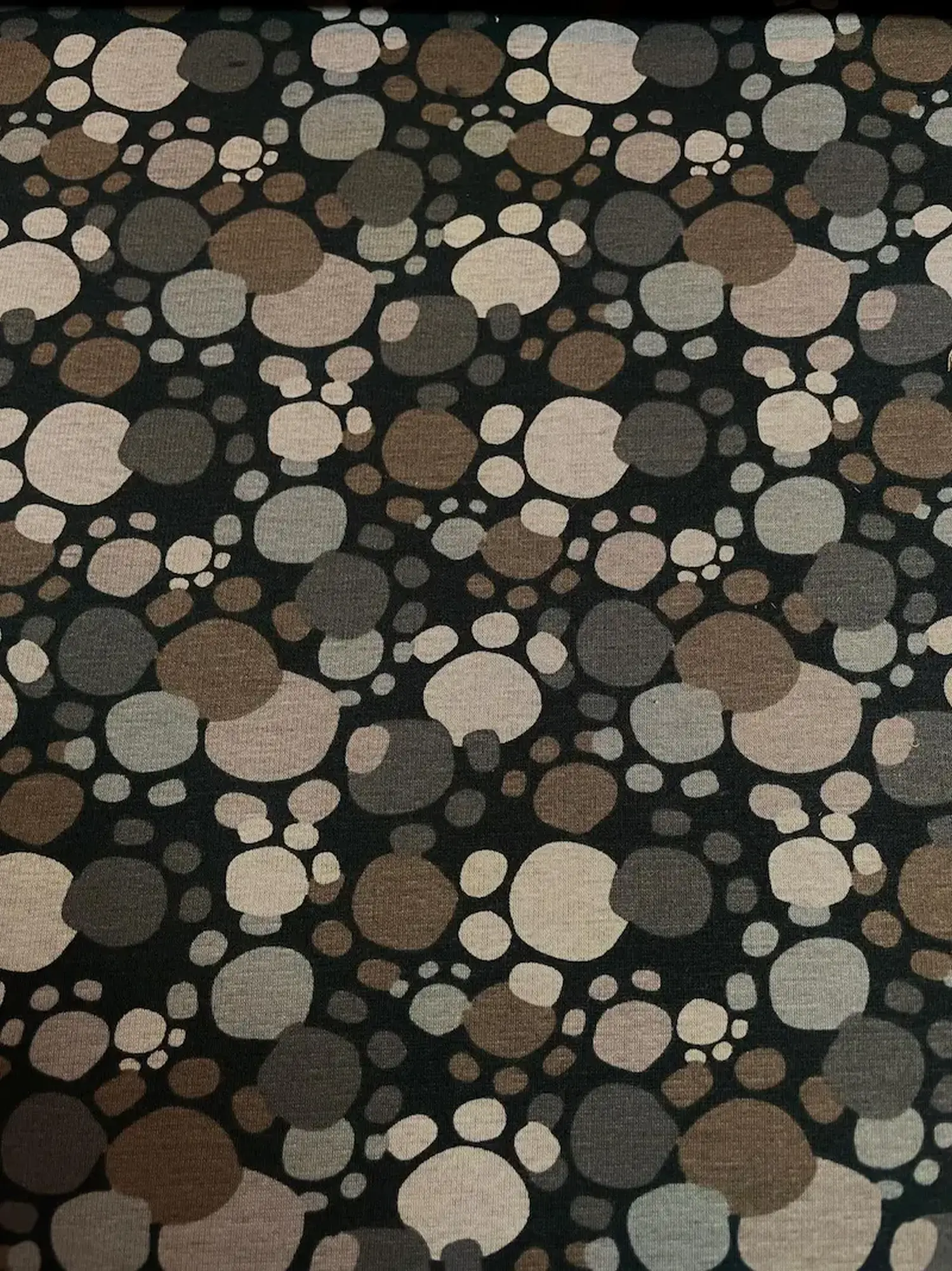 Dunkler Alpenfleece-Stoff mit einem Muster aus abstrakten Pfotenabdrücken und Punkten in verschiedenen Erdtönen wie Braun, Taupe, Beige und Grau auf schwarzem Hintergrund.