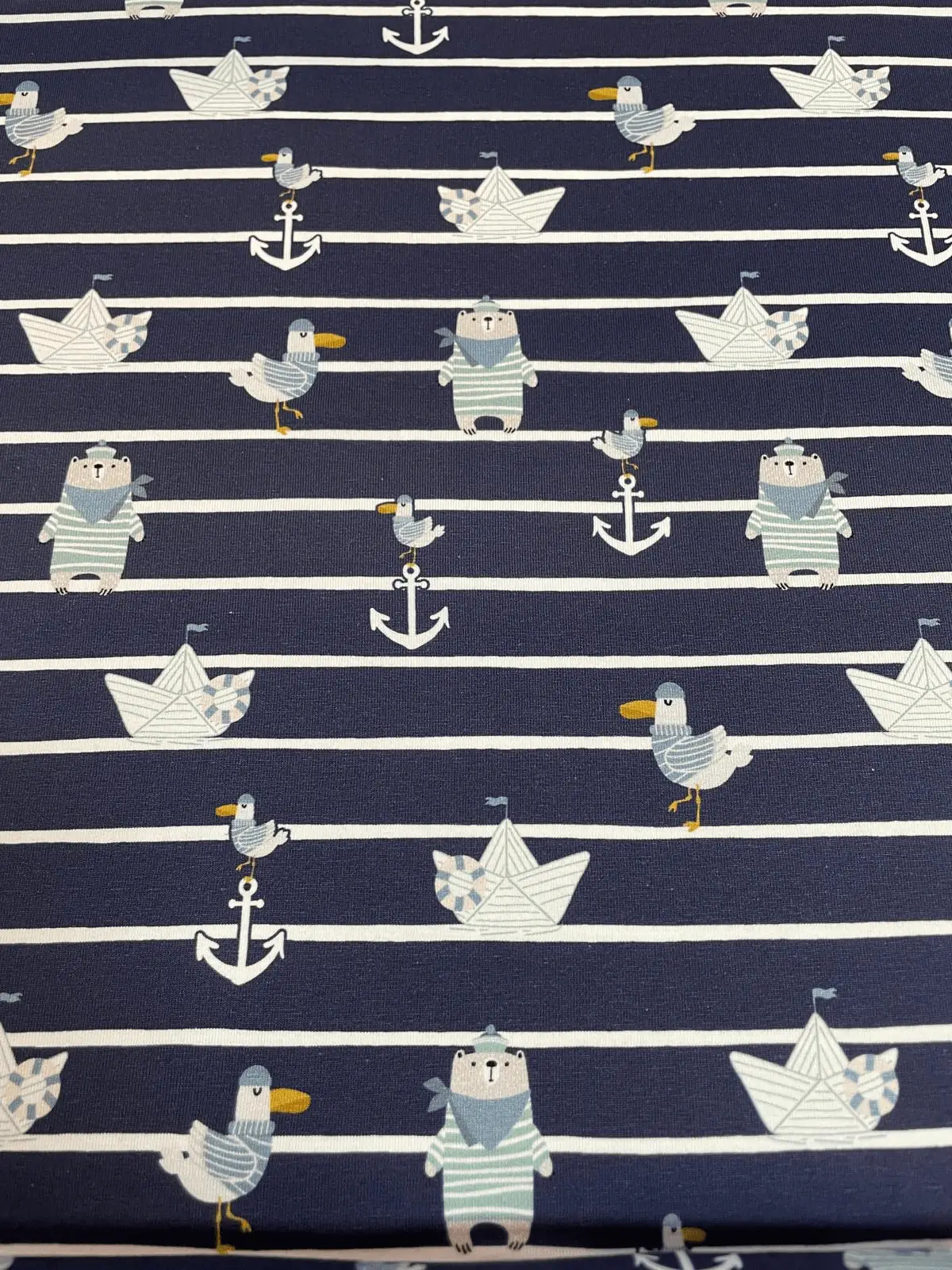 Dunkelblau und weiß quergestreifter Baumwolljersey-Stoff als Meterware, flach fotografiert und bedruckt mit maritimen Kindermotiven wie kleinen Eisbären im Matrosenlook, Möwen, Ankern und Papierschiffchen.