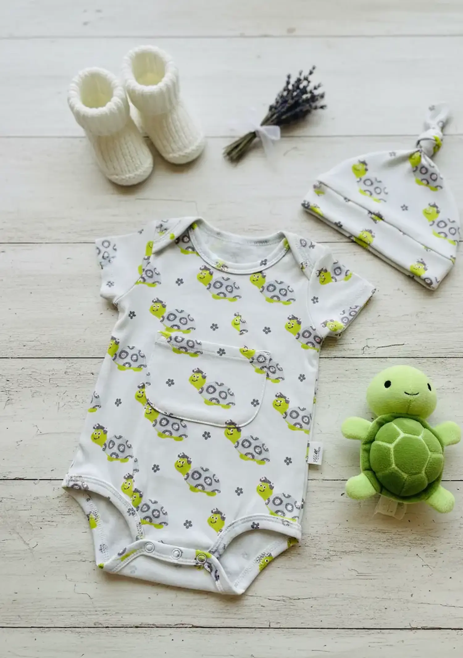 Designbeispiel für Jersey-Stoff: Ein selbstgenähter Baby-Kurzarmbody mit Brusttasche und passendem Knotenmützchen aus hellgrauem Schildkröten-Stoff, dekoriert mit Babyschuhen und einem Plüsch-Schildkrötenspielzeug.