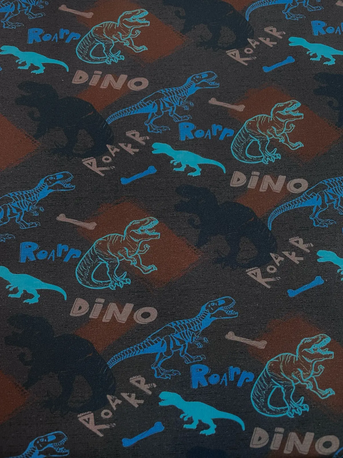 Dunkelblauer Baumwolljersey als Meterware, bedruckt mit einem coolen Dinosaurier-Muster aus T-Rex-Silhouetten, hellblauen Dinos, Knochen und den Schriftzügen Dino und Roar.
