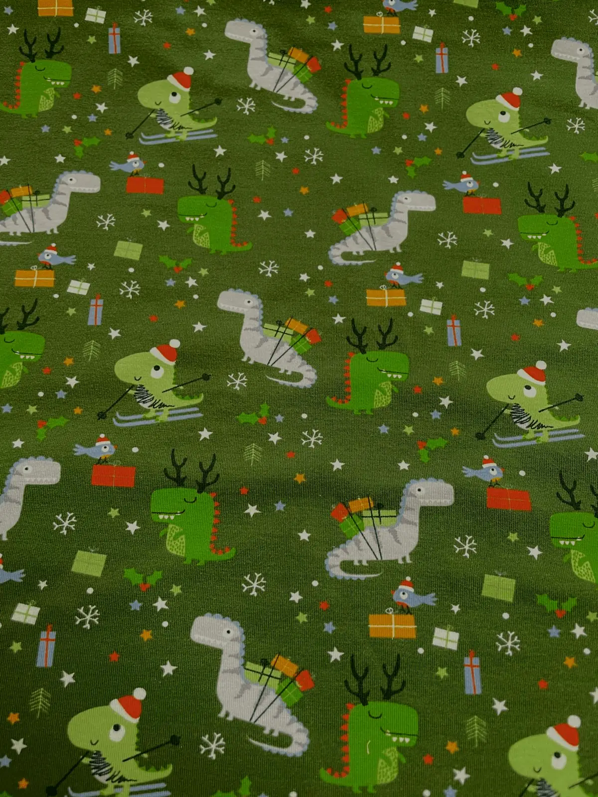 Grüner Baumwoll-Jersey als Meterware mit niedlichem Weihnachts-Dino-Muster (Dinosaurier beim Skifahren, mit Geschenken und Rentiergeweih), Schneeflocken und Sternen.