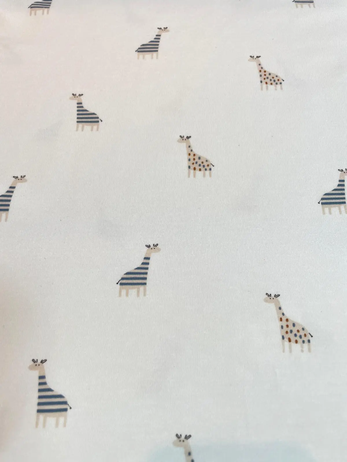 Weißer Jersey-Stoff mit kleinen Giraffen-Motiven, die abwechselnd mit blauen Streifen und ockerfarbenen Punkten gemustert sind.