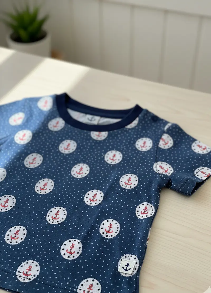 Ein dunkelblaues Kinder-T-Shirt aus Baumwolljersey mit weißen Punkten und roten Anker-Motiven liegt auf einem hellen Tisch. Das Shirt hat ein dunkelblaues Halsbündchen und zeigt das maritime Muster des Stoffes in der Anwendung.