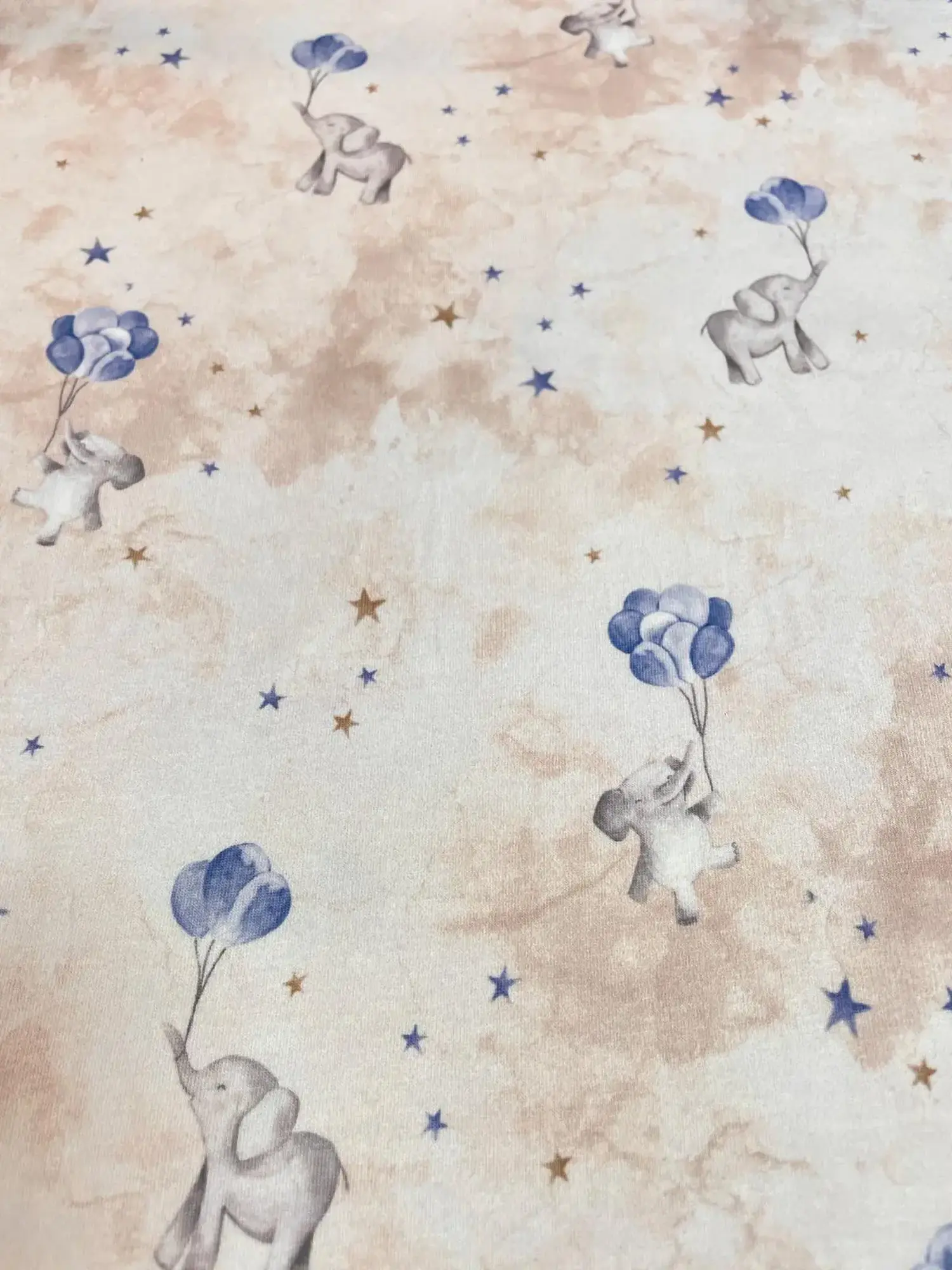 Musteransicht eines creme-beigen Jersey-Stoffs mit Aquarell-Elefanten, blauen Luftballons und kleinen Sternen in Blau und Gold.