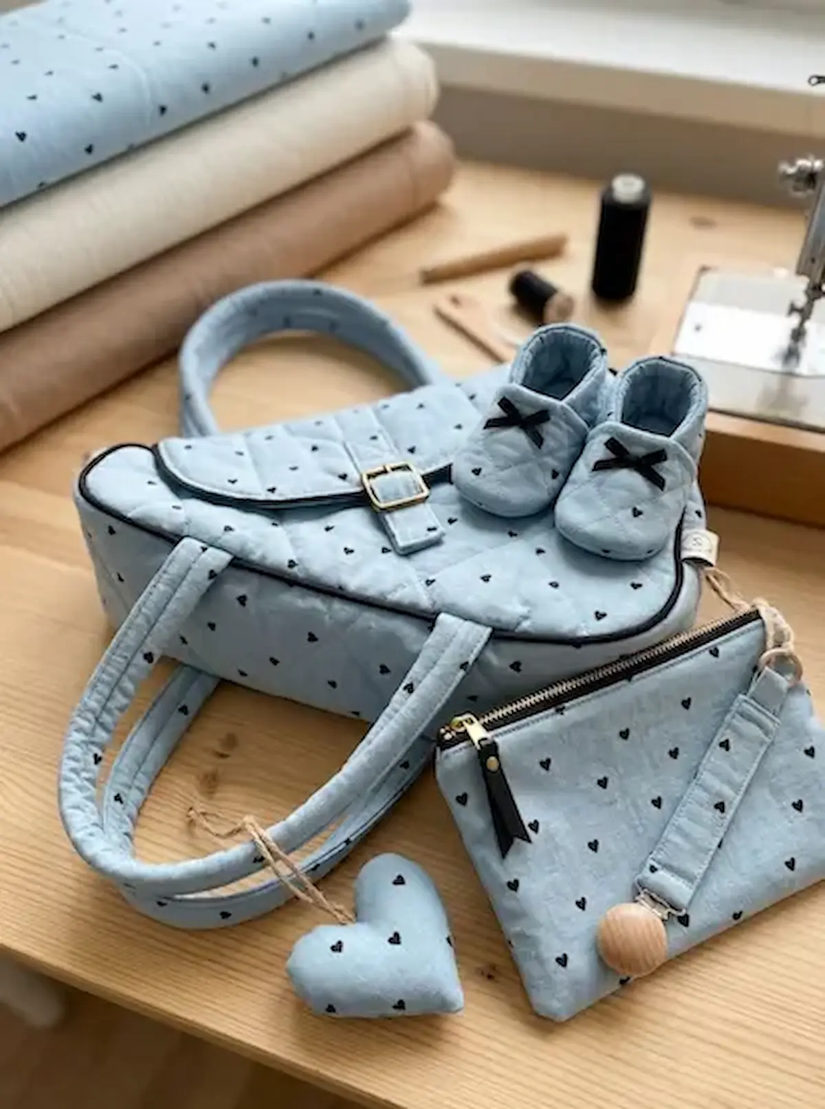 Set aus hellblauer Baumwoll-Webware mit schwarzen Herzen: Gesteppte Tasche, Babyschuhe und kleines Täschchen auf hellem Holzgrund