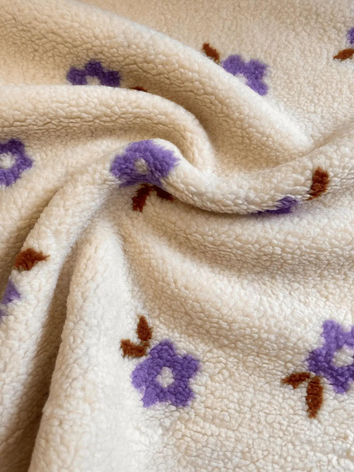 Drapierten Teddy-Plüsch-Stoff als Meterware in Cremeweiß mit violettem Blumenmuster und flauschiger Textur im Detail.