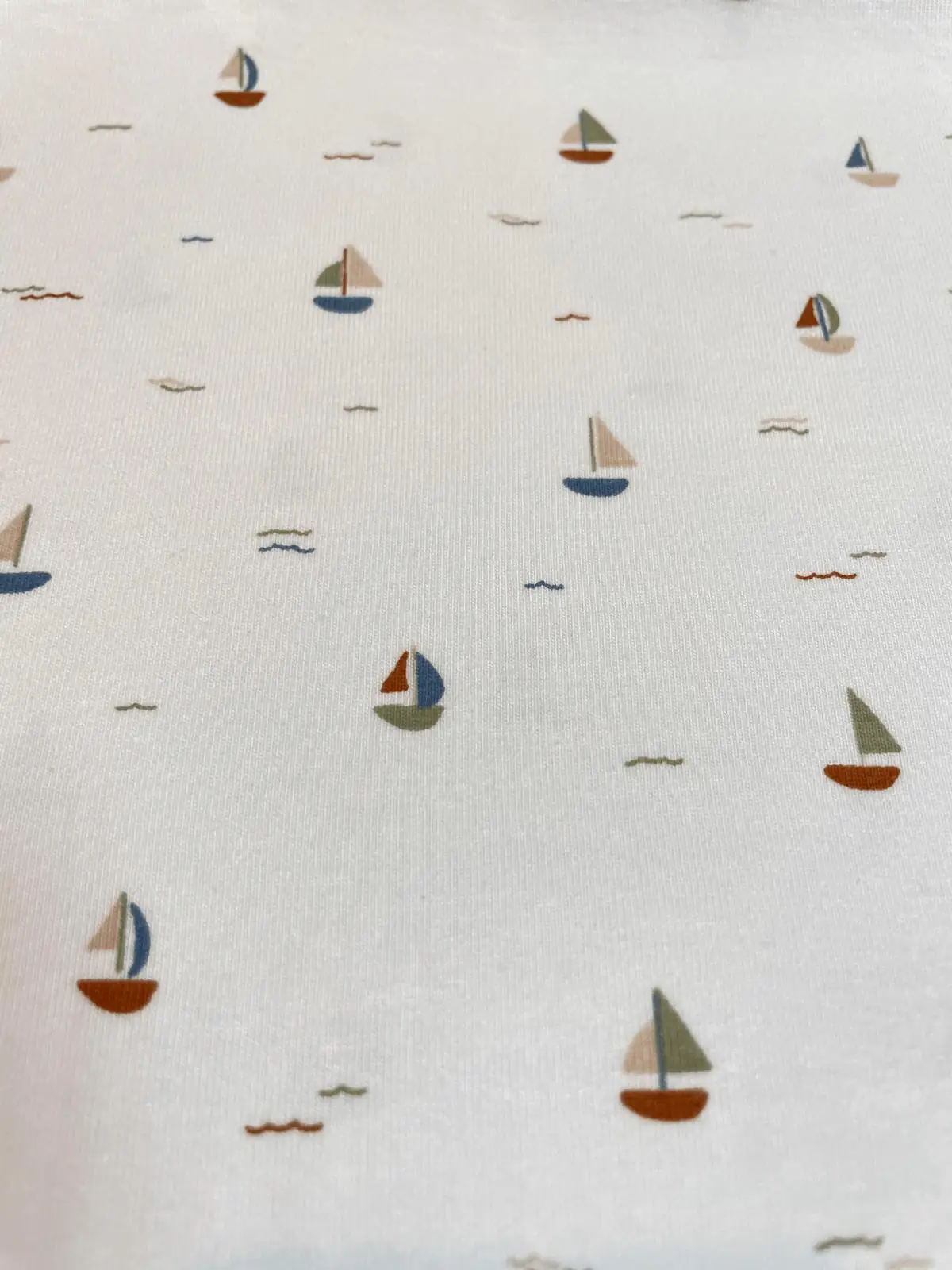 Weißer Jersey-Stoff mit kleinen bunten Segelbooten und dezenten Wellen-Linien in Blau, Rostbraun und Salbeigrün.