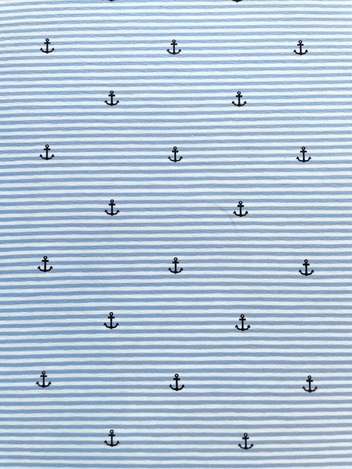Nahaufnahme von elastischem Baumwolljersey in Hellblau und Weiß feingestreift, verziert mit einem regelmäßigen Muster aus kleinen dunkelblauen Ankern; hochwertige Meterware für maritime Babykleidung, T-Shirts und sommerliche Accessoires.