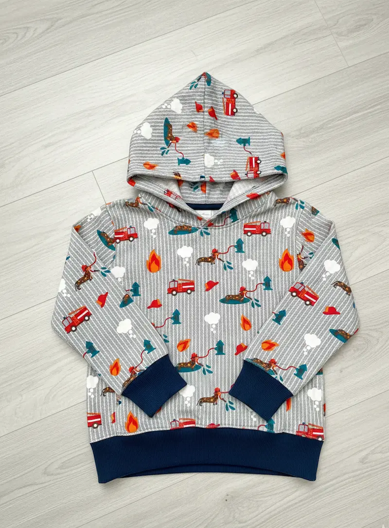 Ein handgenähter Kinder-Kapuzenpullover (Hoodie) flach liegend auf hellem Boden. Der Pullover ist aus hellgrauem, weiß gestreiftem Baumwolljersey mit bunten Feuerwehr-Motiven und Dackeln gefertigt. Die Bündchen an Ärmeln und Saum sind in kontrastreichem D