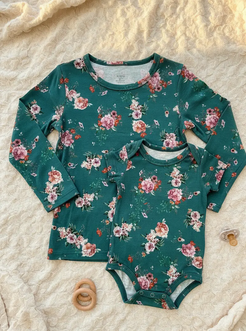 Designbeispiel für Jersey-Stoff Meterware: Ein selbstgenähtes Kinder-Langarmshirt und ein passender Kurzarm-Babybody aus dunkelgrünem Stoff mit Pfingstrosen-Muster liegen flach auf einer beigen Waffelpique-Decke neben einem Holzring und einem Schnuller.