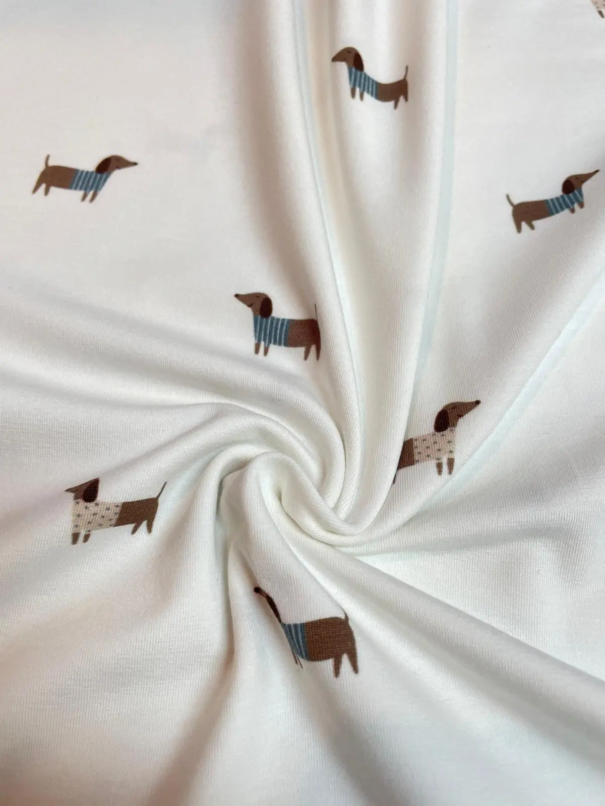 Nahaufnahme von drapiertem Jersey-Stoff mit niedlichem Dackel-Print. Der Stoff ist zu einer Schnecke gelegt, um den weichen, elastischen Faltenwurf zu präsentieren. Der weiße Stoff zeigt die braunen Hunde in gestreiften Pullovern in einer haptischen Detai