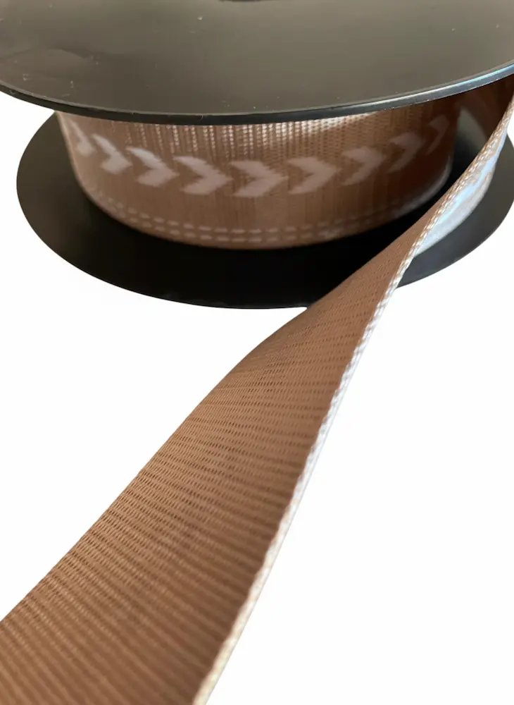 Beigefarbenes Gurtband mit 38mm Breite und eingewebtem weißen Pfeilmuster auf schwarzer Rolle, stabile Meterware für Taschenriemen