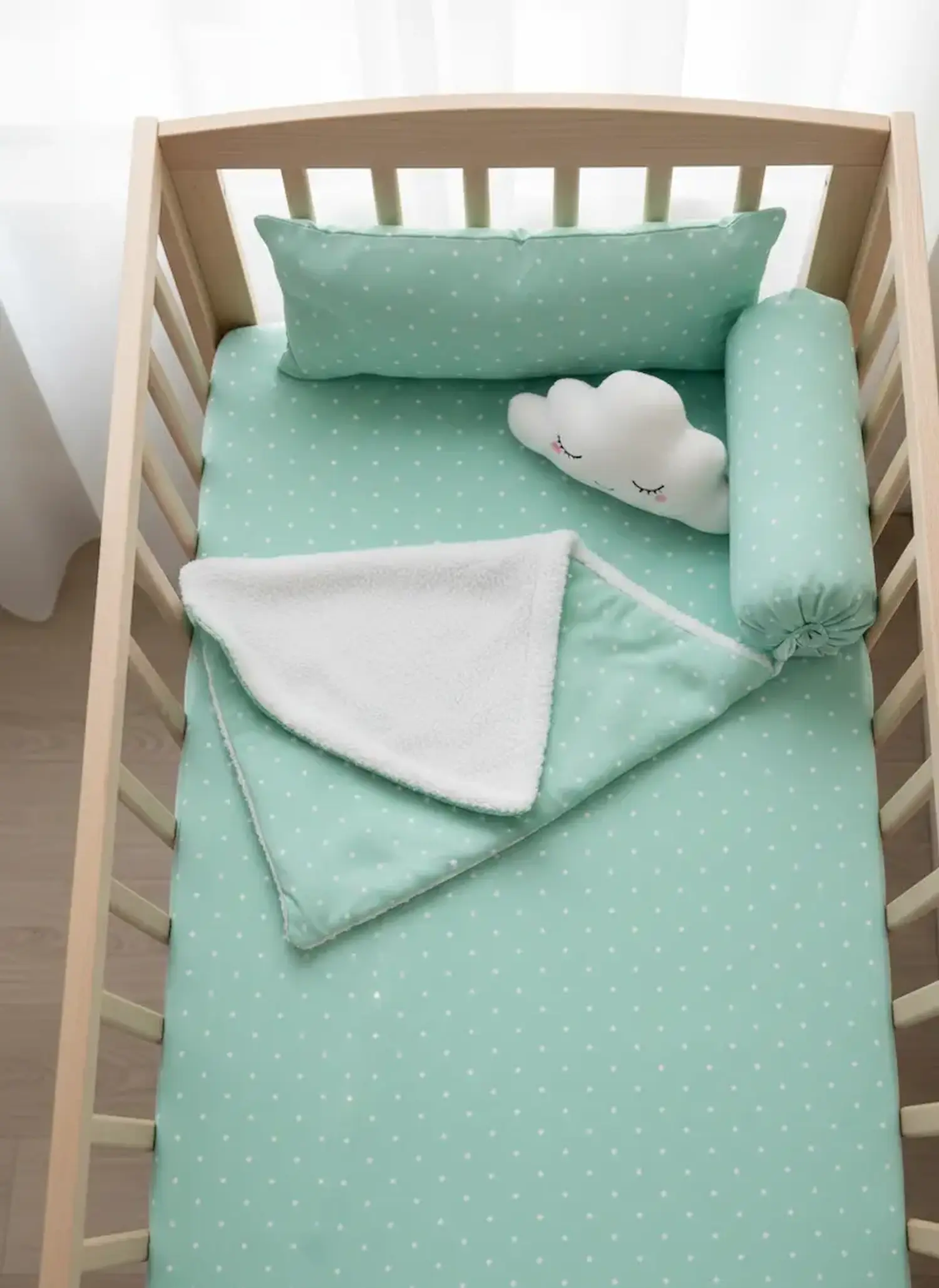 Ein helles Holz-Gitterbett ist mit einem Laken, Kissen und einer Nackenrolle aus mintgrünem Jerseystoff mit kleinen weißen Sternen ausgestattet; eine kuschelige weiße Decke und ein wolkenförmiges Kissen ergänzen das Set.