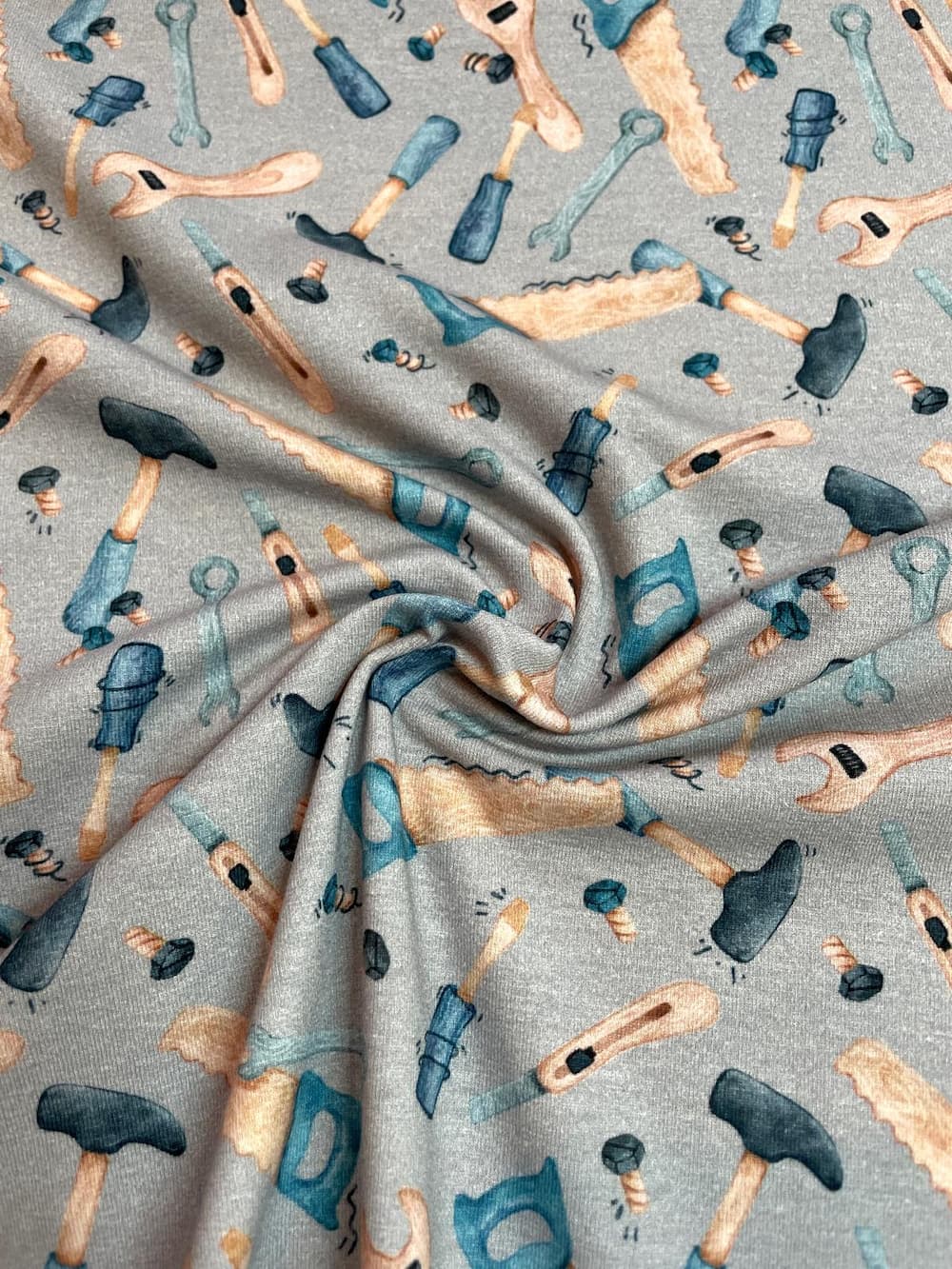 Ein kunstvoll drapierter, hellgrau melierter Jerseystoff als Meterware, bedruckt mit einem bunten Werkzeug-Muster aus Hämmern, Sägen, Schraubenschlüsseln, Schraubendrehern und Schrauben.