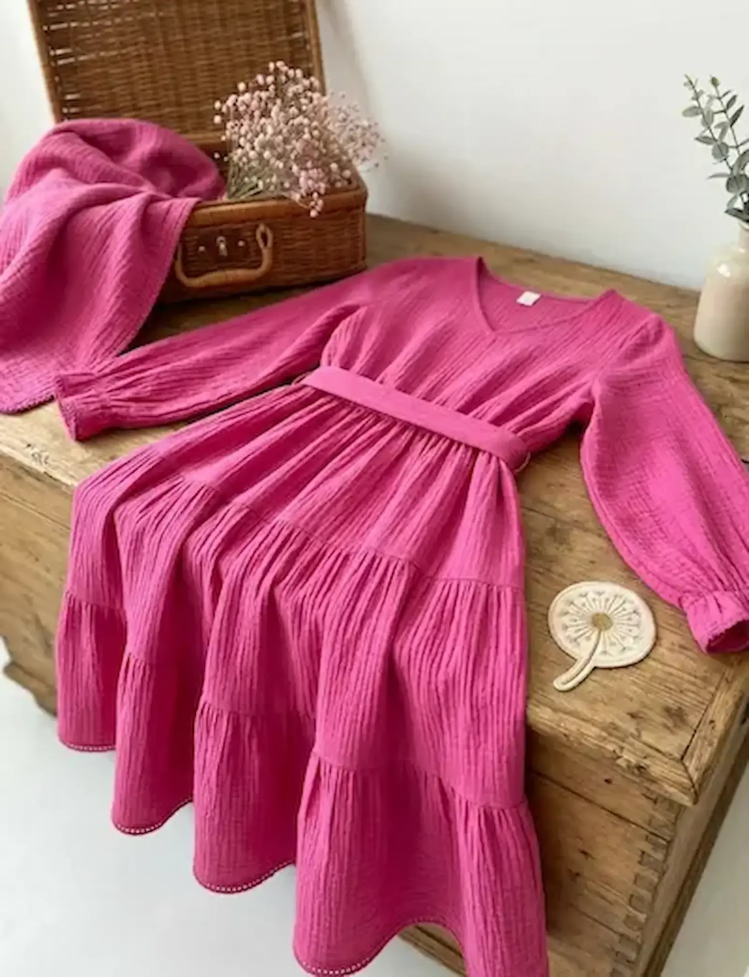 Langärmeliges Stufenkleid aus pinkem Musselin Stoff auf einer Holzkiste, farbenfrohe Double Gauze Baumwoll-Meterware für Damen- und Kinderkleidung.