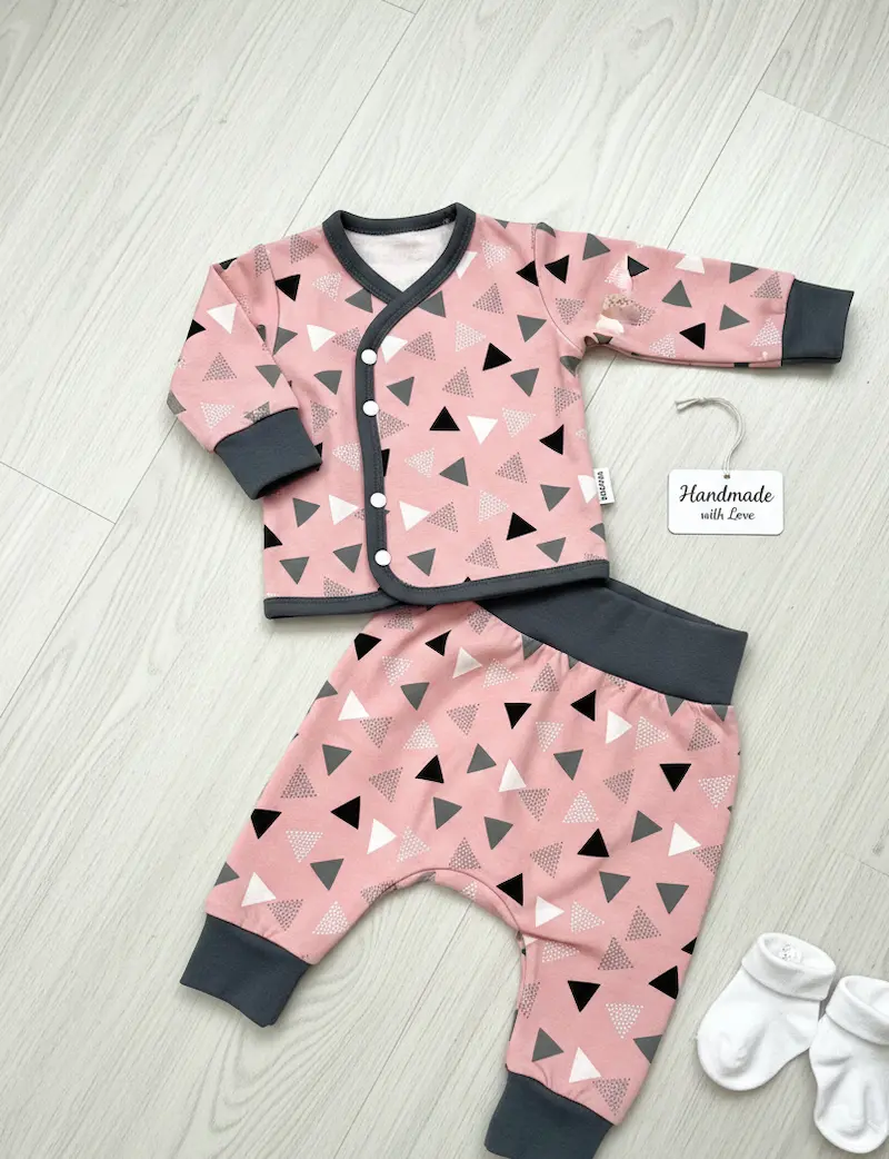 Designbeispiel aus unserem Baumwolljersey mit Dreiecken in Rosa: Ein handgenähtes Baby-Set bestehend aus einer langärmligen Wickeljacke mit Druckknöpfen und einer passenden Pumphose, abgesetzt mit dunkelgrauen Bündchen. Daneben liegt ein Schild mit der Au