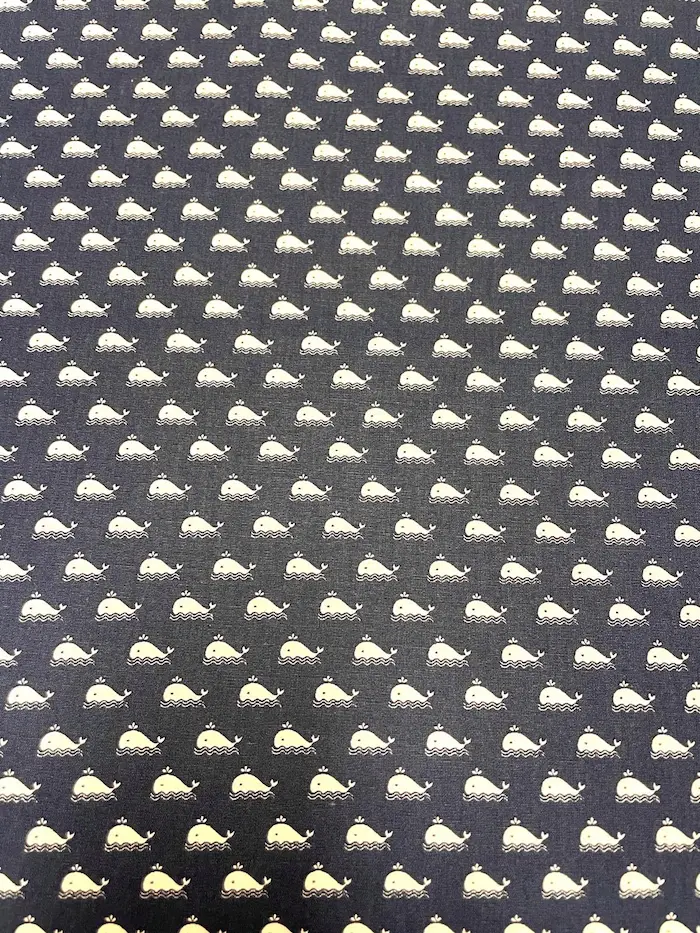 Eine flächendeckende Draufsicht auf unelastische Baumwoll-Popeline in Dunkelblau, bedruckt mit einem feinen, regelmäßigen Muster aus kleinen weißen Walen, die auf minimalistischen Wellen schwimmen.