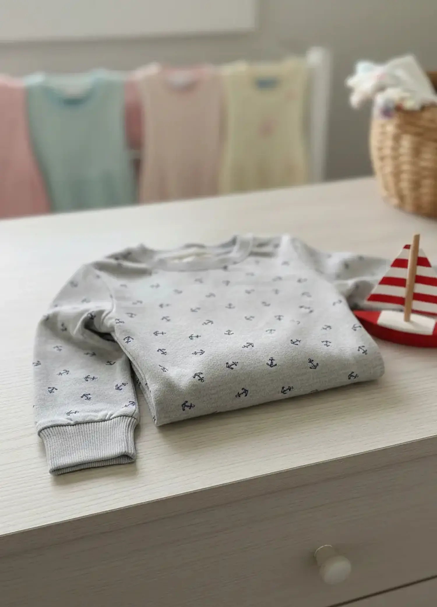 Ein ordentlich gefalteter Kinderpullover aus hellgrauem Jerseystoff mit kleinen dunkelblauen Ankern liegt auf einer hellen Kommode neben einem kleinen Holzsegelboot.