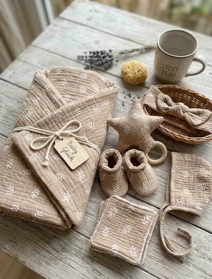 Hochwertige Musselin-Webware in Beige mit weißem Blätter-Print, verarbeitet zu einem Baby-Geschenkset mit Decke, Schühchen, Mütze und Rassel.