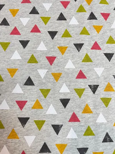 Grau-melierter Sweatstoff mit einem gleichmäßigen Muster aus bunten Dreiecken in den Farben Senfgelb, Rostrot, Olivgrün, Weiß und Anthrazit.