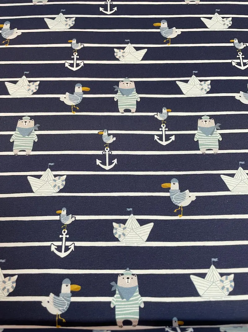 Dunkelblau und weiß quergestreifter Baumwolljersey-Stoff als Meterware, flach fotografiert und bedruckt mit maritimen Kindermotiven wie kleinen Eisbären im Matrosenlook, Möwen, Ankern und Papierschiffchen.