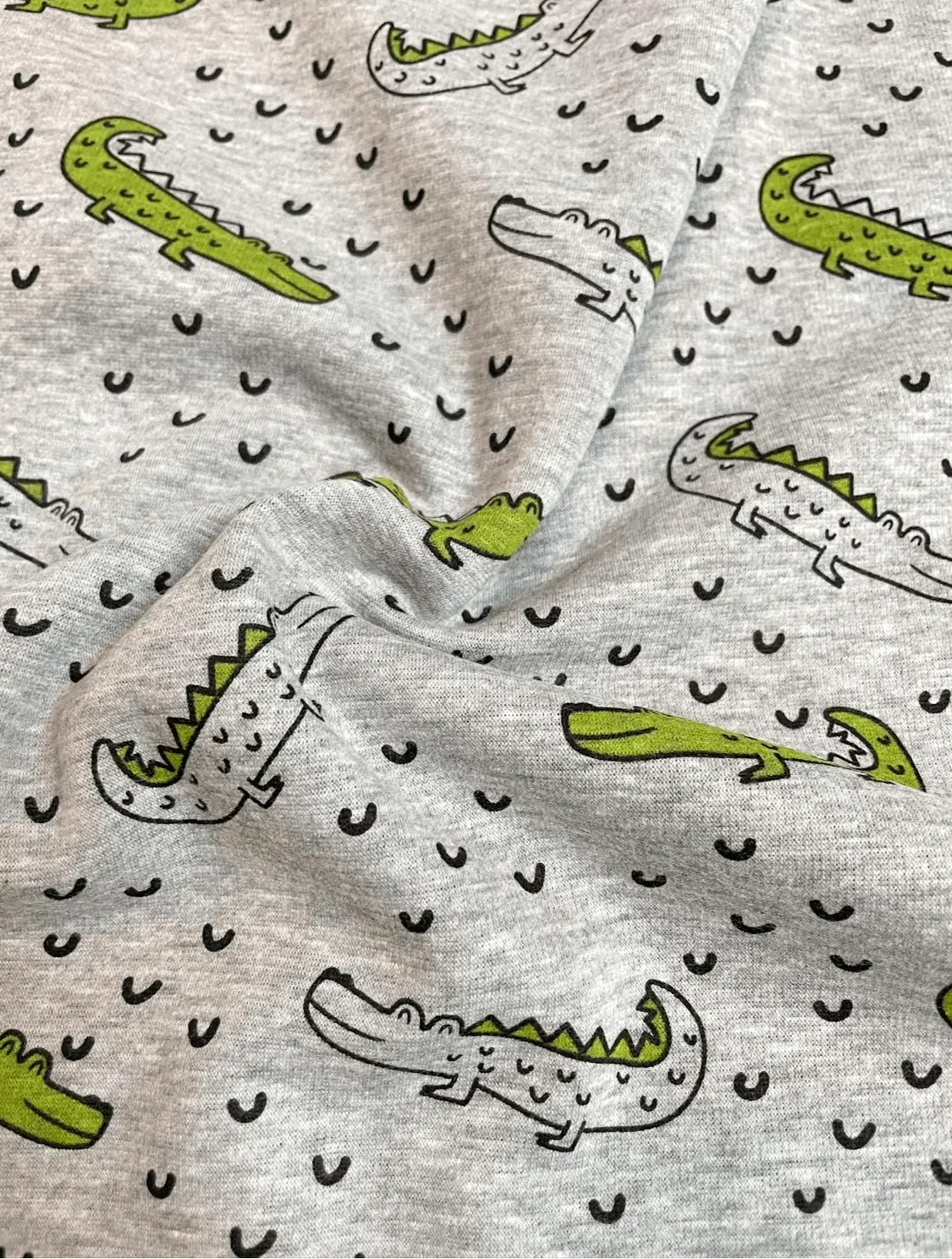 Detailaufnahme eines weichen Alpenfleece-Stoffes in Grau-Melange mit Krokodil-Motiven. Die Stofffalten zeigen die geschmeidige, schwere Qualität des Materials für Winterbekleidung.
