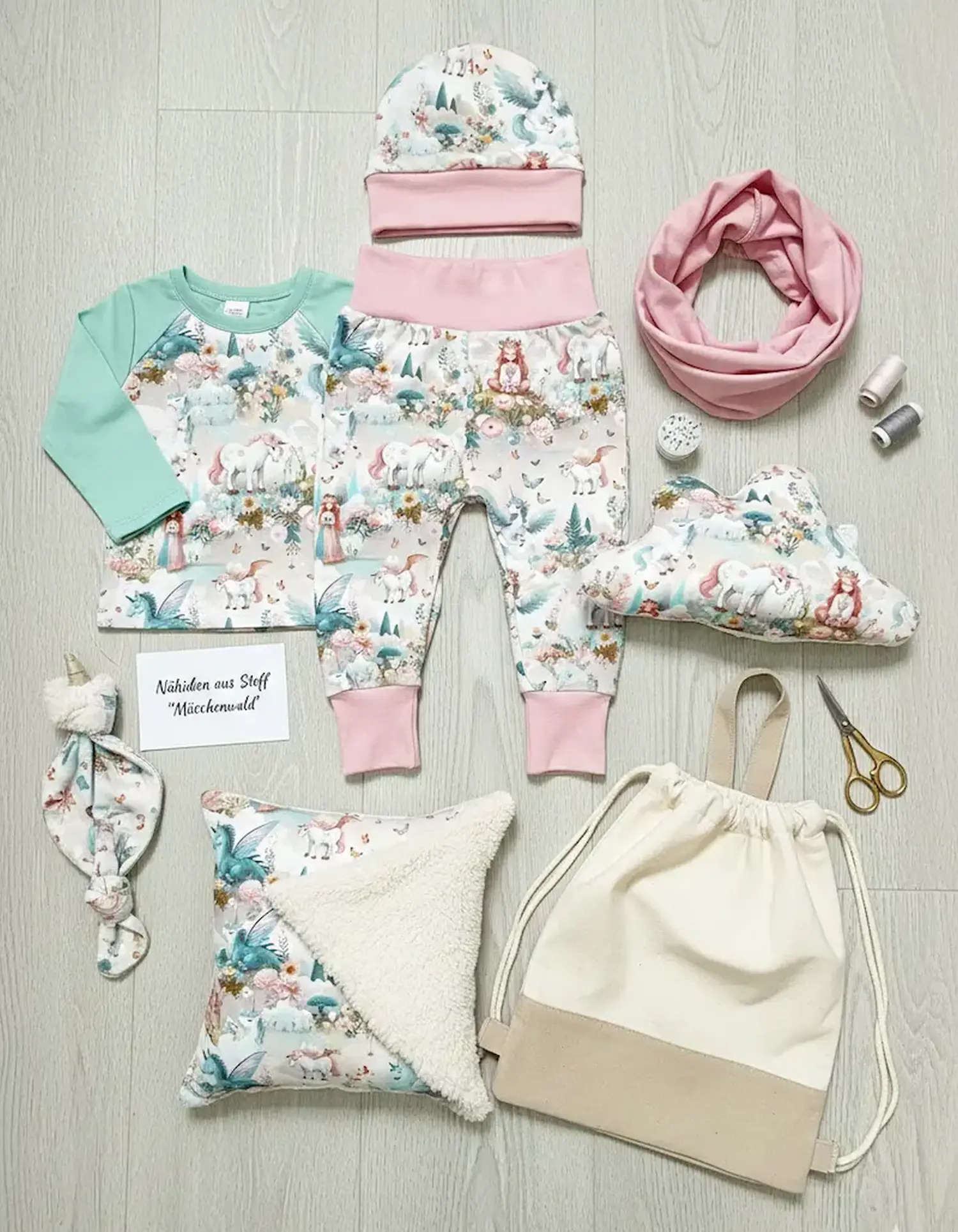 Flachgereihte Nähideen aus Kinder-Jerseystoff mit Einhorn-, Feen- und Prinzessinnen-Motiven: Babyhose, Raglanshirt mit mintgrünen Ärmeln, Mütze, Loop-Schal, Wolkenkissen und ein Schnuffeltuch auf Holzboden.