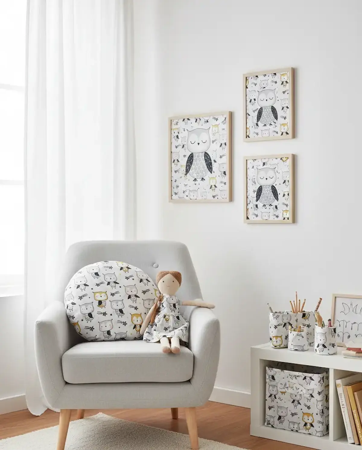 Ein helles, skandinavisch eingerichtetes Kinderzimmer mit einem grauen Sessel, auf dem ein rundes Patchwork-Kissen mit Eulen-Motiv und eine Stoffpuppe liegen. An der Wand hängen drei passende Bilderrahmen und auf dem Regal stehen Stoffkörbchen aus demselb