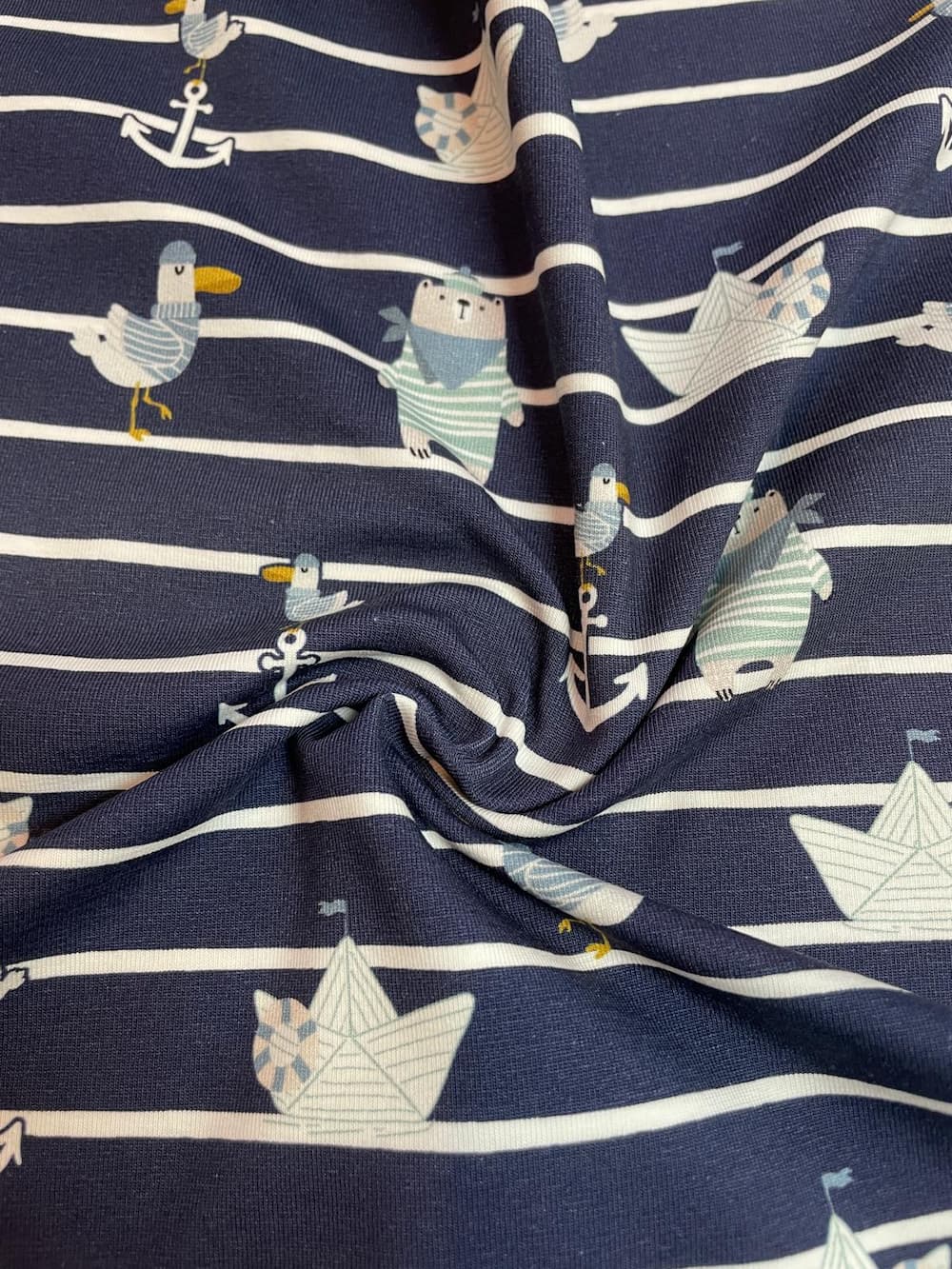 Ein kunstvoll drapierter Baumwolljersey-Stoff als Meterware mit dunkelblauen und weißen Querstreifen. Durch den weichen Faltenwurf kommen die kleinen, gedruckten Motive wie Bären im Matrosenlook, Möwen und Papierschiffchen schön zur Geltung.