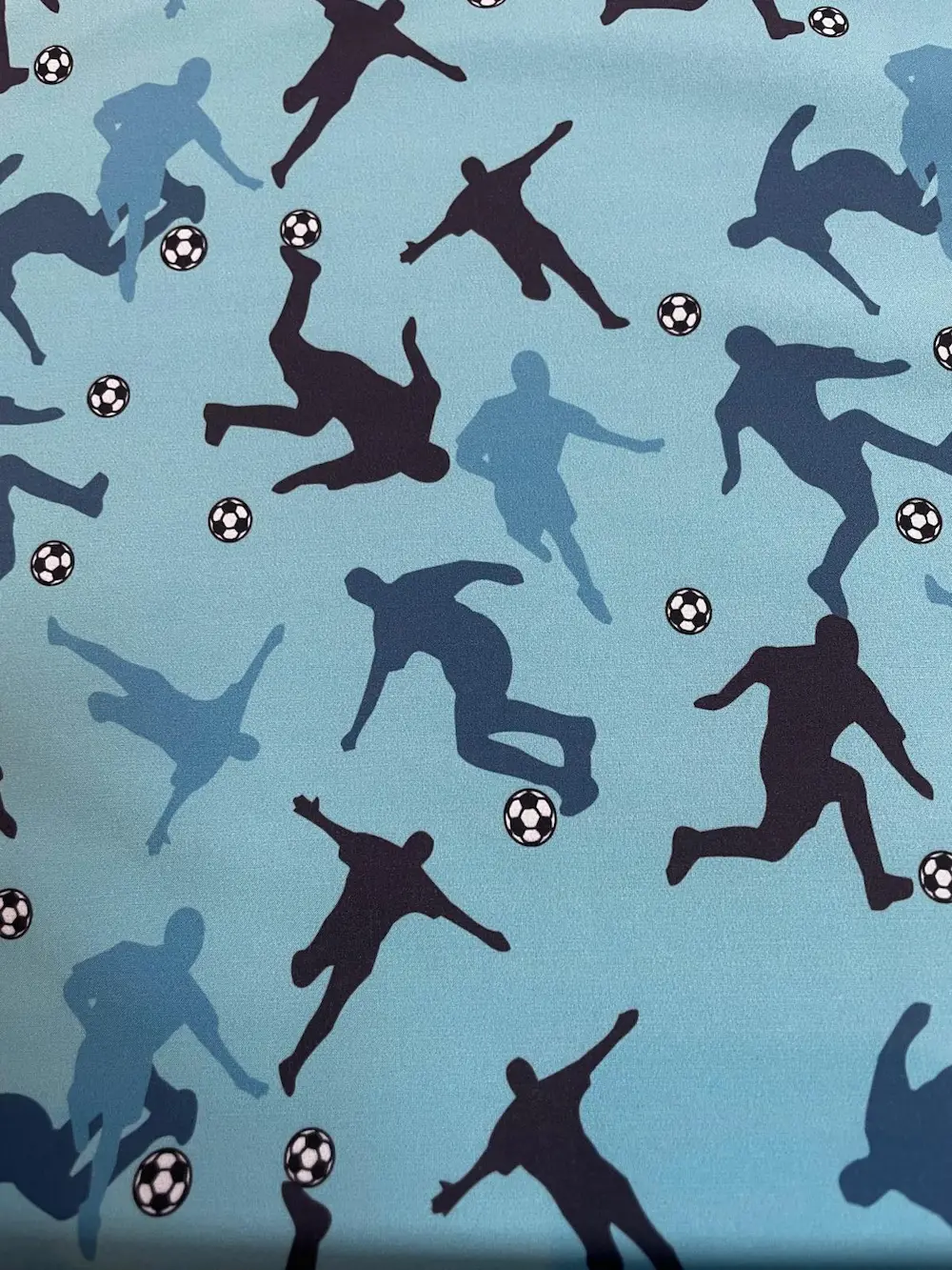 Hellblauer Softshell-Stoff mit einem sportlichen Fußball-Muster. Zu sehen sind Silhouetten von Fußballspielern in verschiedenen Blau-Nuancen und Schwarz, die in Action-Posen (Dribbeln, Schießen) dargestellt werden. Ergänzt wird das Design durch kleine kla