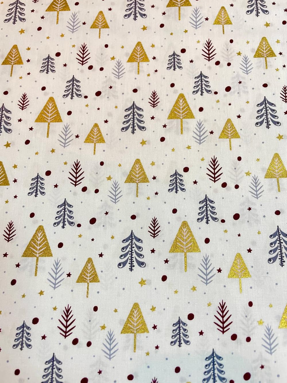 Festlicher Baumwollstoff als Meterware mit einem Muster aus bunten Tannenbäumen in glitzerndem Gold, Dunkelrot und Grau sowie kleinen Sternen und Punkten auf hellem weißem Hintergrund.