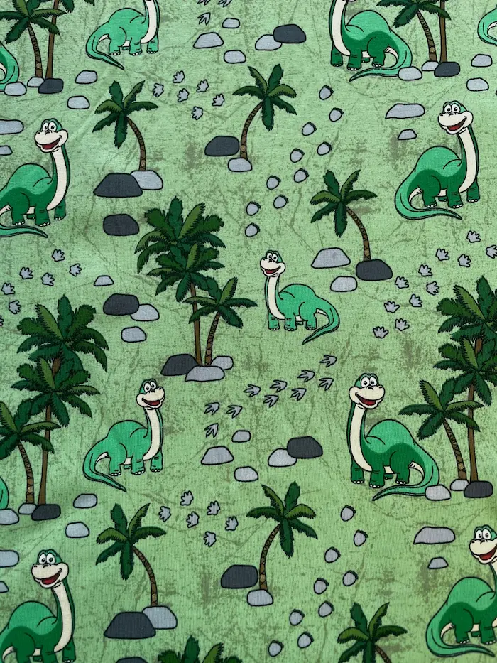 Hochwertiger Baumwolljersey als Meterware in Hellgrün mit einem fröhlichen Dinosaurier-Muster. Zu sehen sind freundliche, grüne Langhals-Dinos zwischen tropischen Palmen, grauen Steinen und kleinen Fußspuren im Allover-Design.