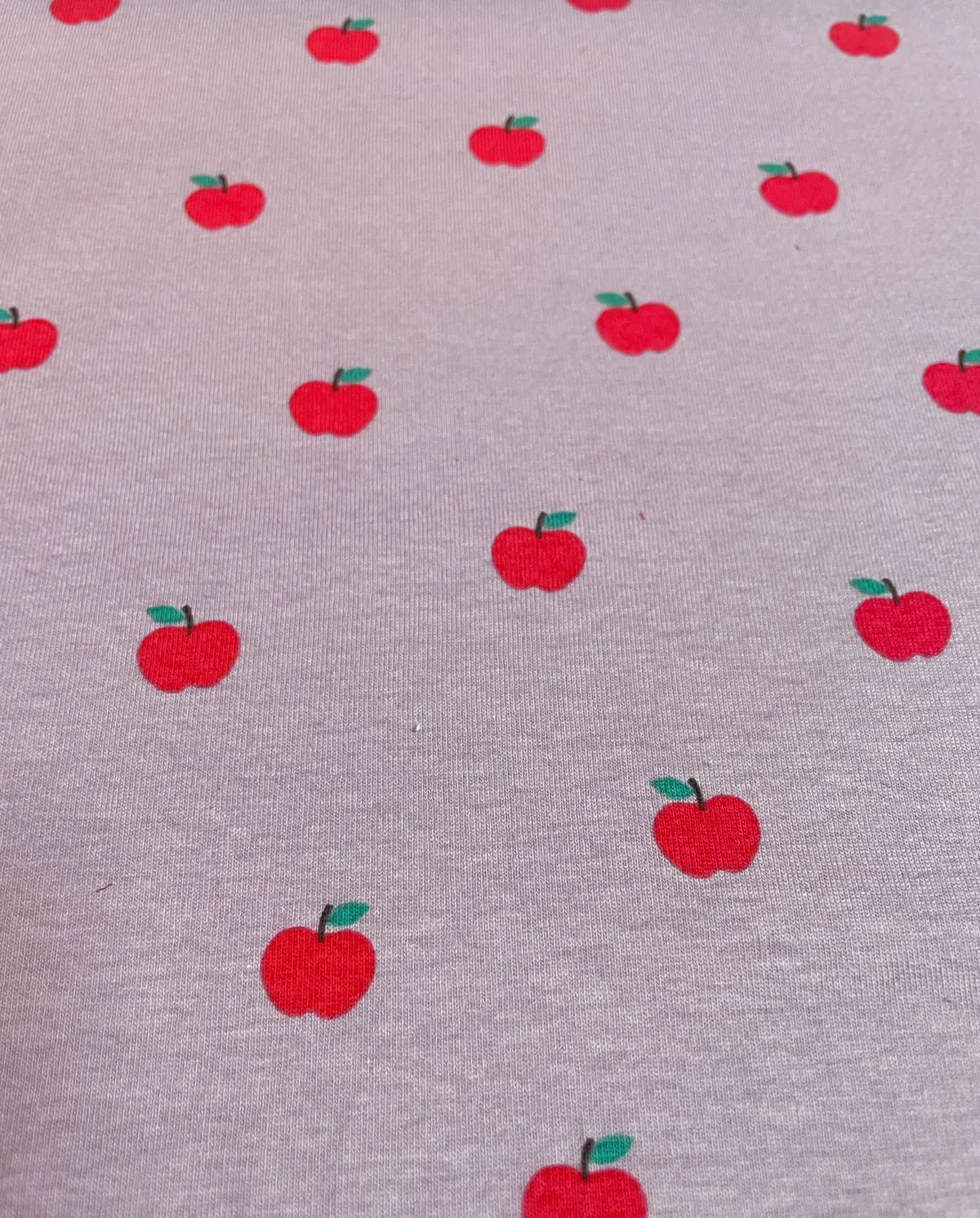 Elastischer Jersey-Stoff in meliertem Altrosa mit einem gleichmäßigen Muster aus kleinen, leuchtend roten Äpfeln mit grünen Blättern. Ideal für Kinderbekleidung und Accessoires.
