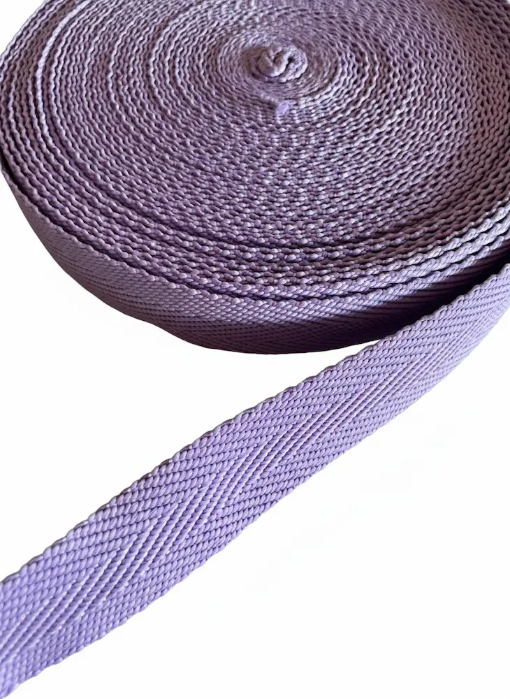 Eine Rolle flieder-lila Gurtband mit 3 cm Breite und Fischgrat-Muster vor weißem Hintergrund, im Vordergrund ein ausgerollter Gurtbandstreifen als Meterware.