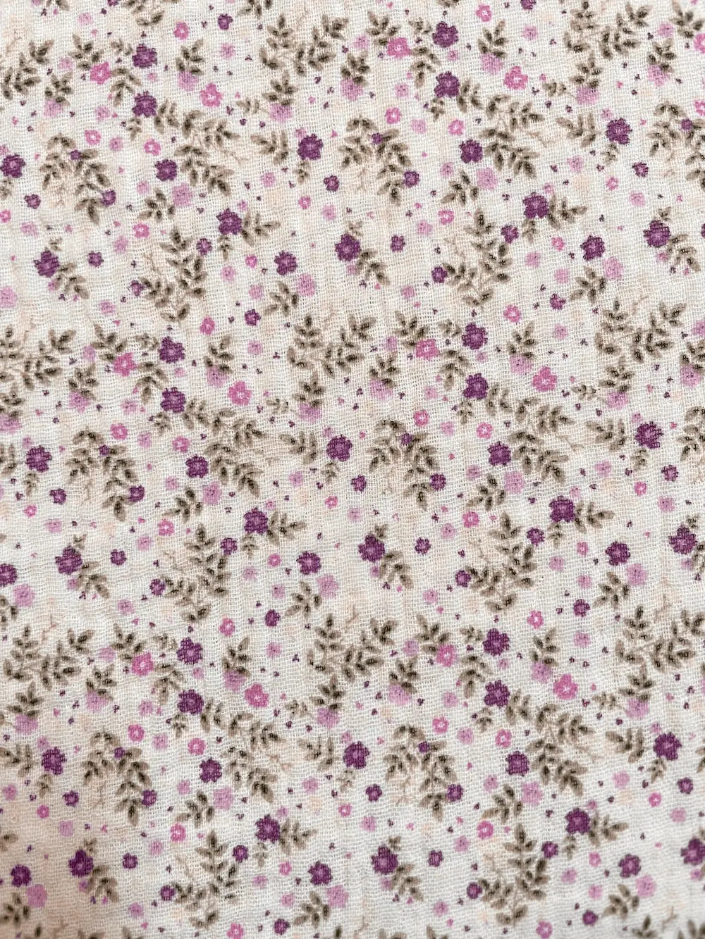 Nahaufnahme von weißem Musselin-Stoff mit kleinen lila Blumen und grünen Blättern, weiche Double-Gauze Struktur aus 100% Baumwolle.