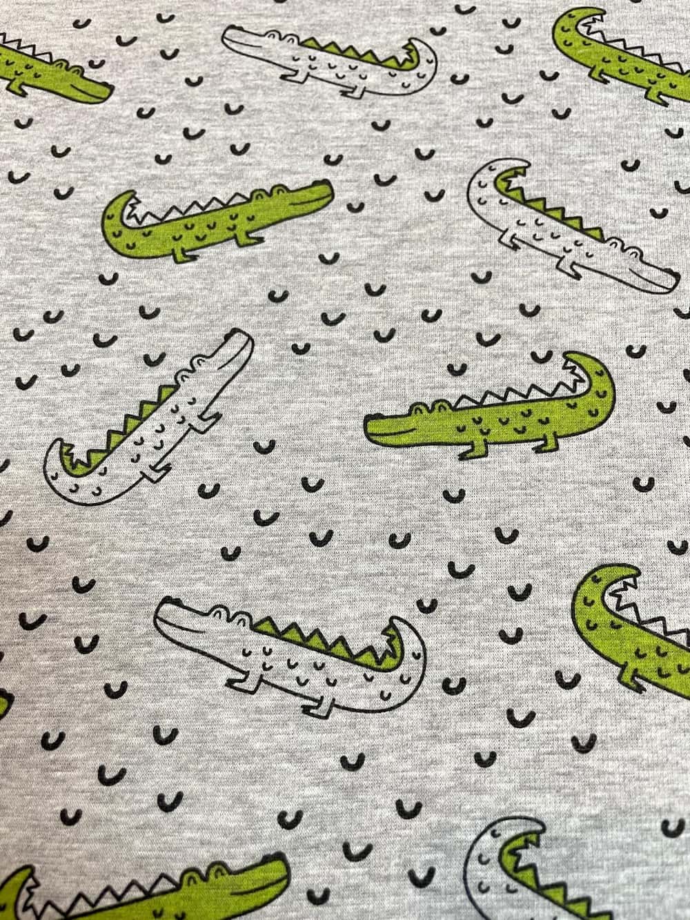 Kuscheliger Alpenfleece-Stoff in Grau-Melange mit Krokodil-Print. Niedliches Reptilien-Muster mit grünen Krokodilen und kleinen Wellen, ideal für warme Kinderkleidung und Decken.