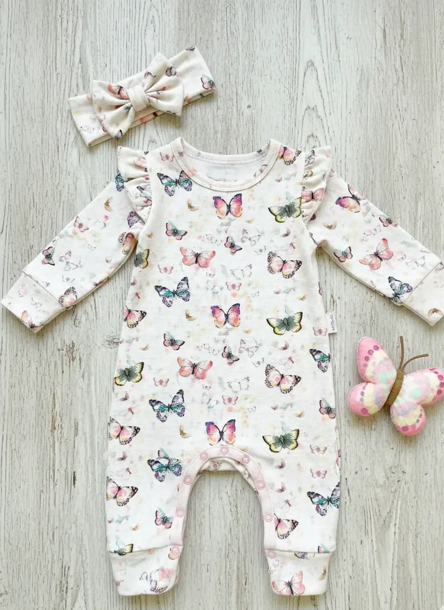 Ein cremefarbener Baby-Strampler aus Modal French Terry mit bunten Aquarell-Schmetterlingen. Das Kleidungsstück liegt flach auf hellem Holz und zeigt Details wie Schulterrüschen und farblich passende Accessoires (Stirnband und Stoffschmetterling).