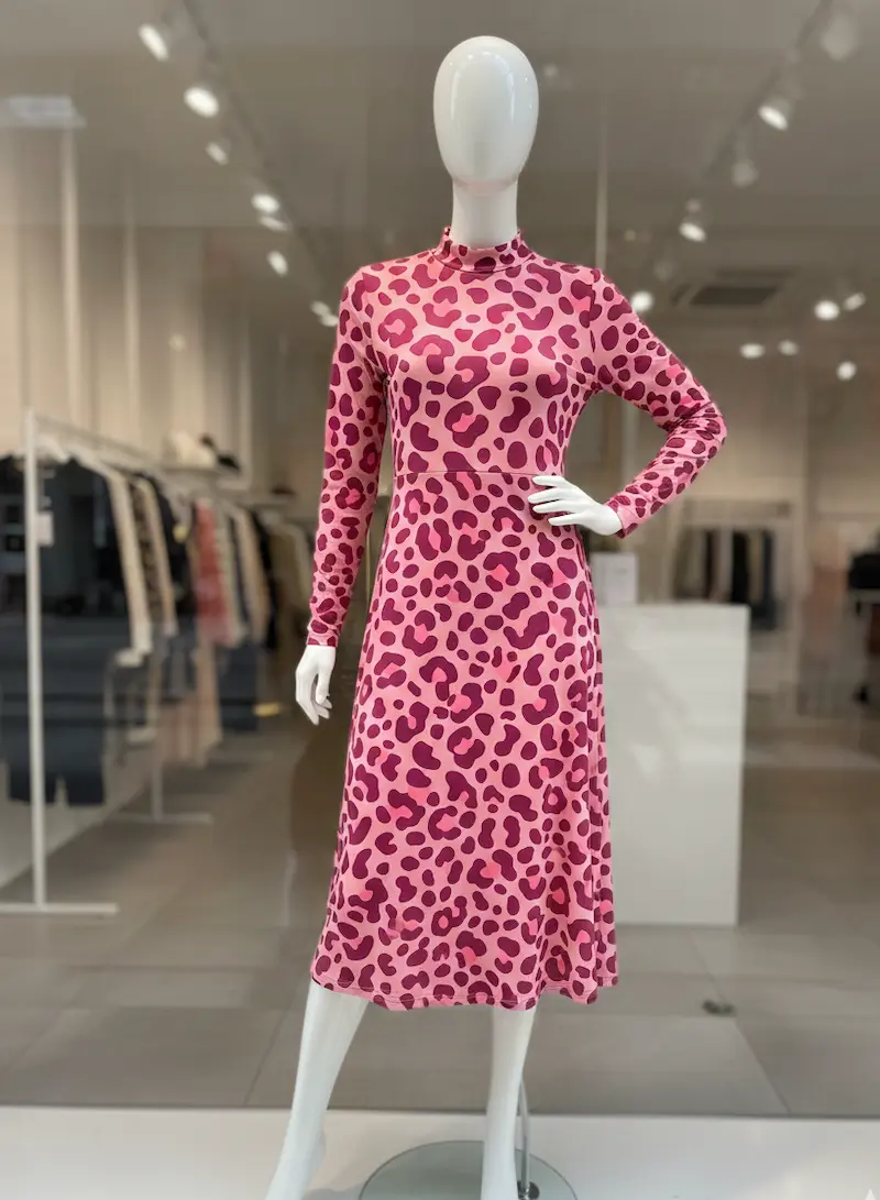 Ein elegantes, langärmliges Midikleid mit Stehkragen, genäht aus weichem Baumwolljersey-Stoff mit auffälligem Leopardenmuster. Das Kleid im Allover-Leo-Design in Pink, Rosa und Beere wird auf einer weißen Schaufensterpuppe in einem hellen Modegeschäft prä