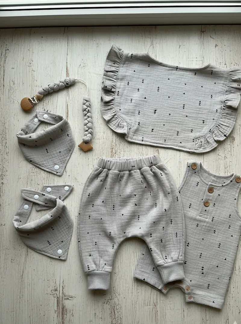 Ein liebevoll gestaltetes, mehrteiliges Baby-Bekleidungsset flach ausgelegt auf hellem Holzboden. Alle Teile, darunter eine gemütliche Pumphose, ein ärmelloser Strampler mit Holzknöpfen, ein Rüschen-Oberteil, zwei Halstücher und eine geflochtene Schnuller