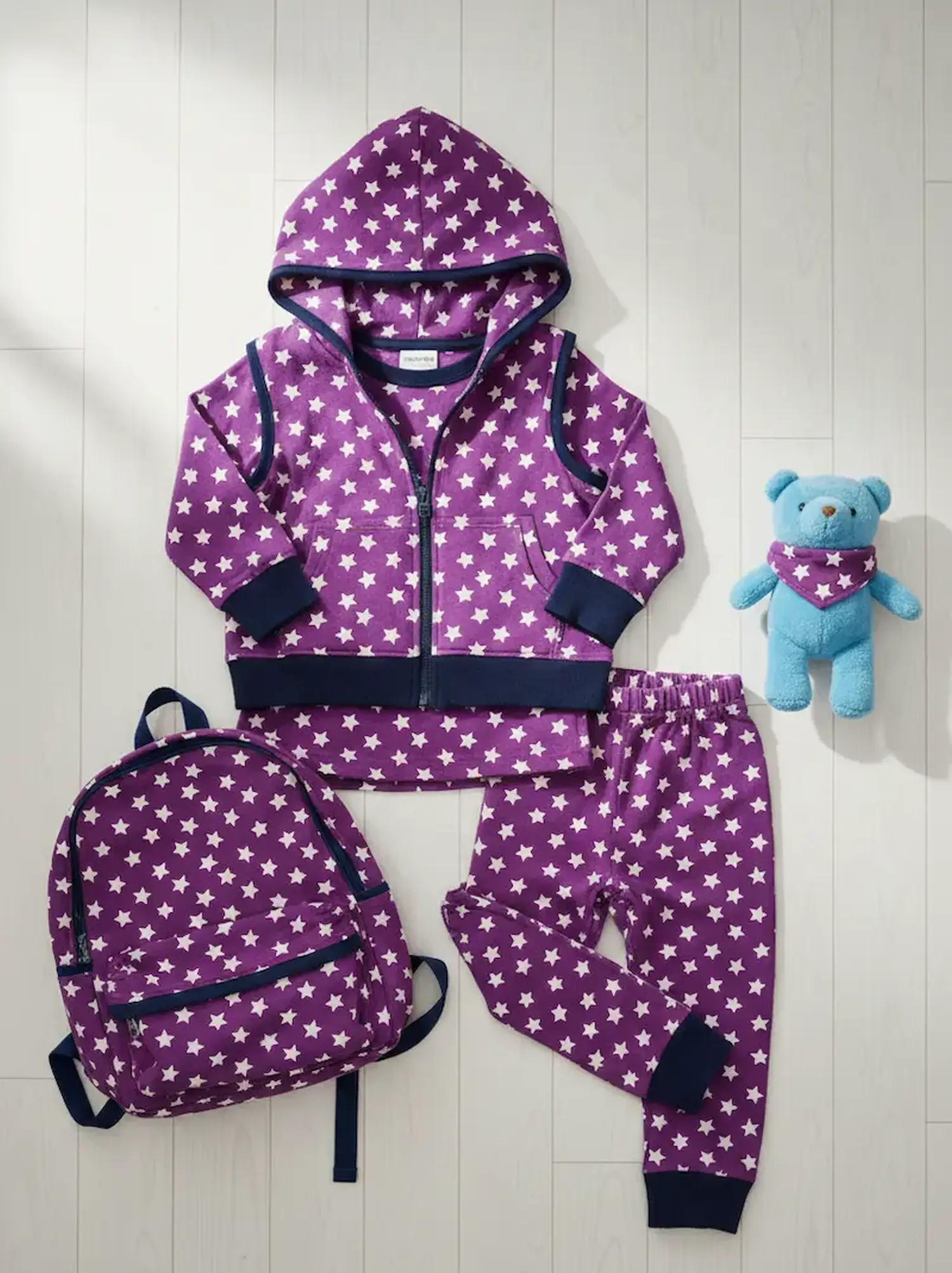 Ein handgenähtes Kinder-Set aus violettem Jerseystoff mit weißen Sternen, bestehend aus einer Kapuzenjacke, einer Hose und einem passenden Rucksack, flach liegend auf hellem Boden neben einem blauen Teddybären.