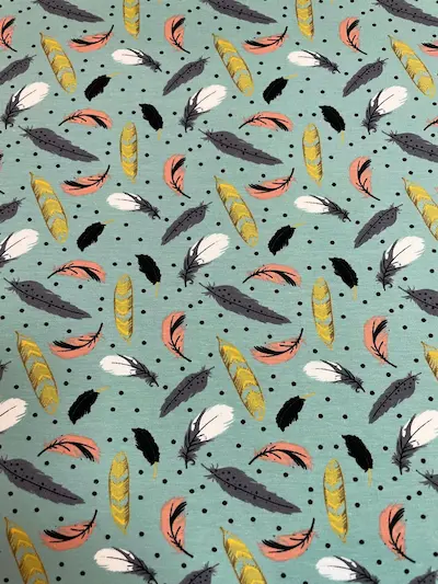 Heller mintgrüner Jersey-Stoff mit einem verspielten Muster aus bunten Federn in Senfgelb, Altrosa, Weiß und Schwarz. Ergänzt wird das Design durch kleine schwarze Pünktchen im Hintergrund, ideal für Baby- und Kinderkleidung im Boho-Stil.