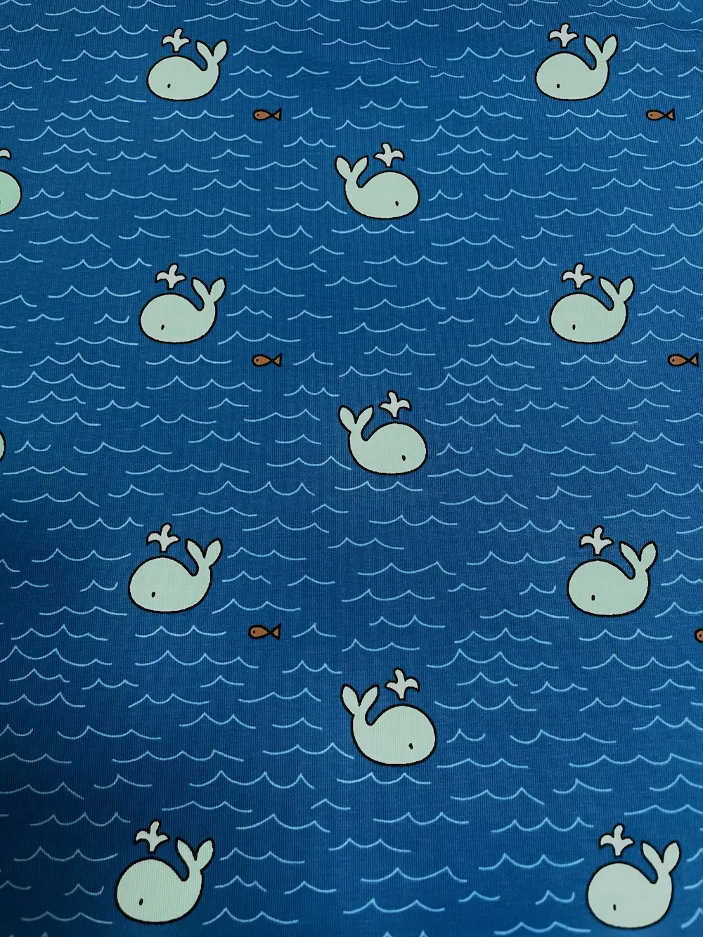 Ein flach liegender Baumwolljersey-Stoff als Meterware in hellem Dunkelblau, bedruckt mit einem maritimen Allover-Muster aus kleinen, hellblauen Walen mit Wasserfontänen, winzigen orangefarbenen Fischen und feinen, hellblauen Wellenlinien.